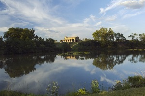 A view across a lake to a house on a hill with the sun shining on it and trees all around